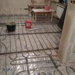 R.P.S-Studio - Montaż ogrzewania podłogowego: rury grzewcze rozłożone na folii z nadrukiem 'ITAL' na podłożu przed wylaniem wylewki, widoczny pojemnik z zaprawą i czerwony kanister w tle, surowe ściany.