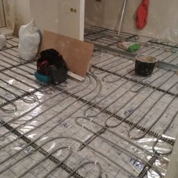 R.P.S-Studio - Montaż ogrzewania podłogowego: rury grzewcze ułożone na folii z nadrukiem, widoczne torby budowlane, wiadro i narzędzia w surowym wnętrzu.