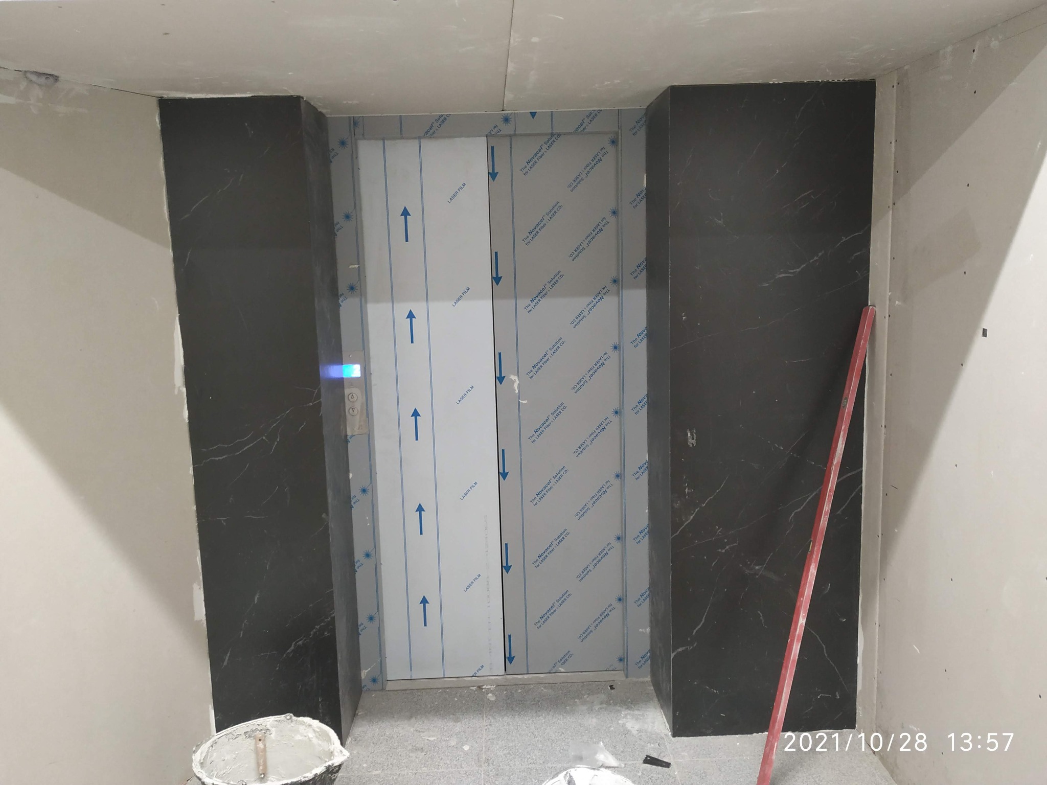 Montaż obudowy windy z czarnego marmuru, zabezpieczona folią ochronną, widoczny poziomica i wiadro z zaprawą.