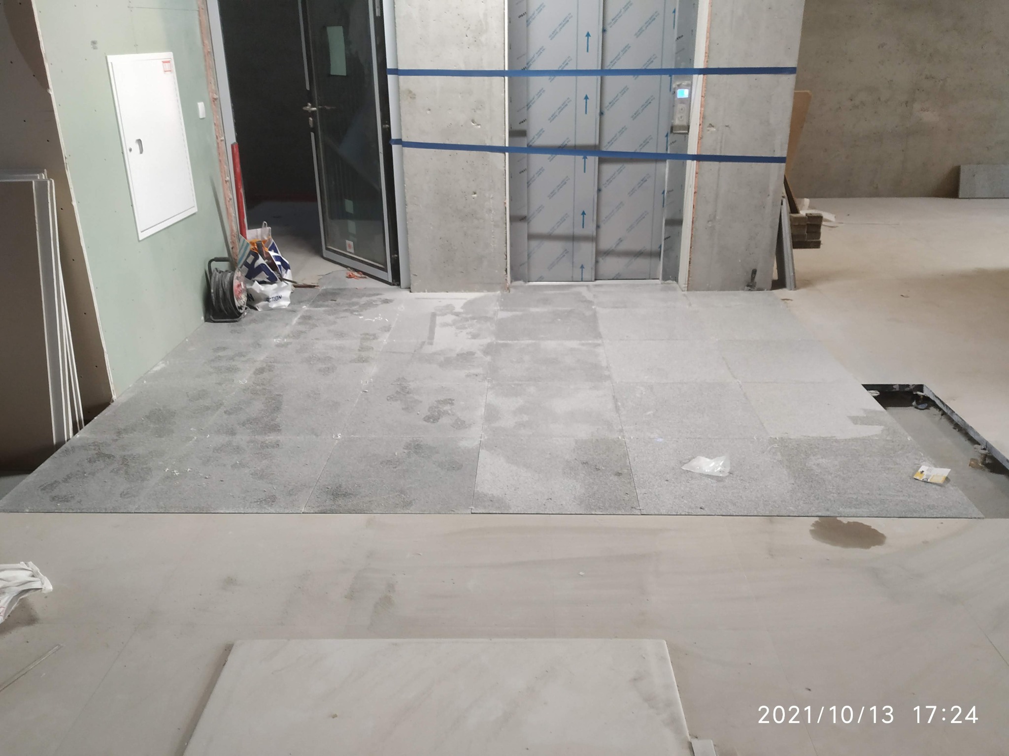 Montaż szarych płytek podłogowych w nowym budynku z widoczną windą i ścianą z płyt kartonowo-gipsowych, częściowo wykończona podłoga, widoczne niedociągnięcia i brakujące fragmenty płytek.