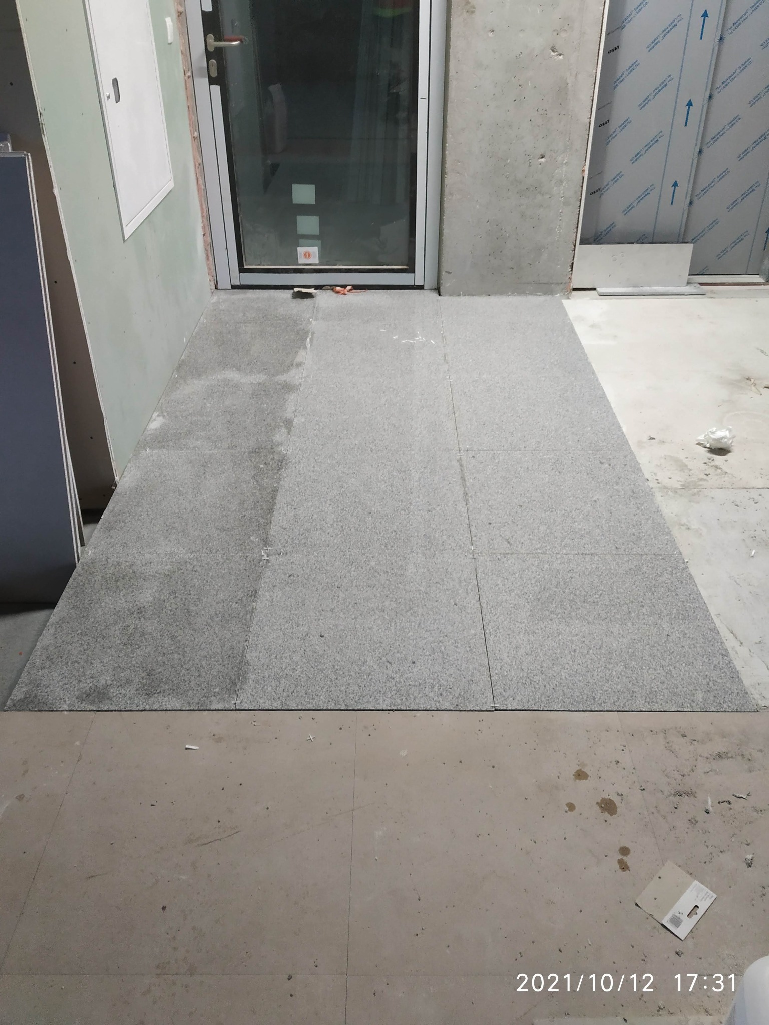 Układanie szarych płytek podłogowych w przedpokoju, widoczne nierówności podłoża i resztki po remoncie.