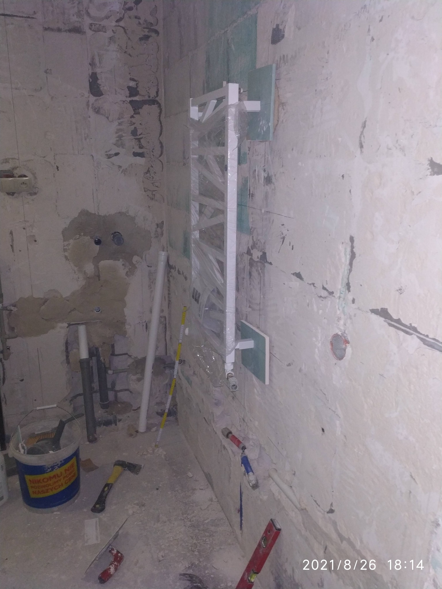 Łazienka w trakcie remontu: surowe ściany z widocznymi instalacjami, grzejnik drabinkowy przymocowany do ściany, wiadro z narzędziami i poziomica na podłodze.