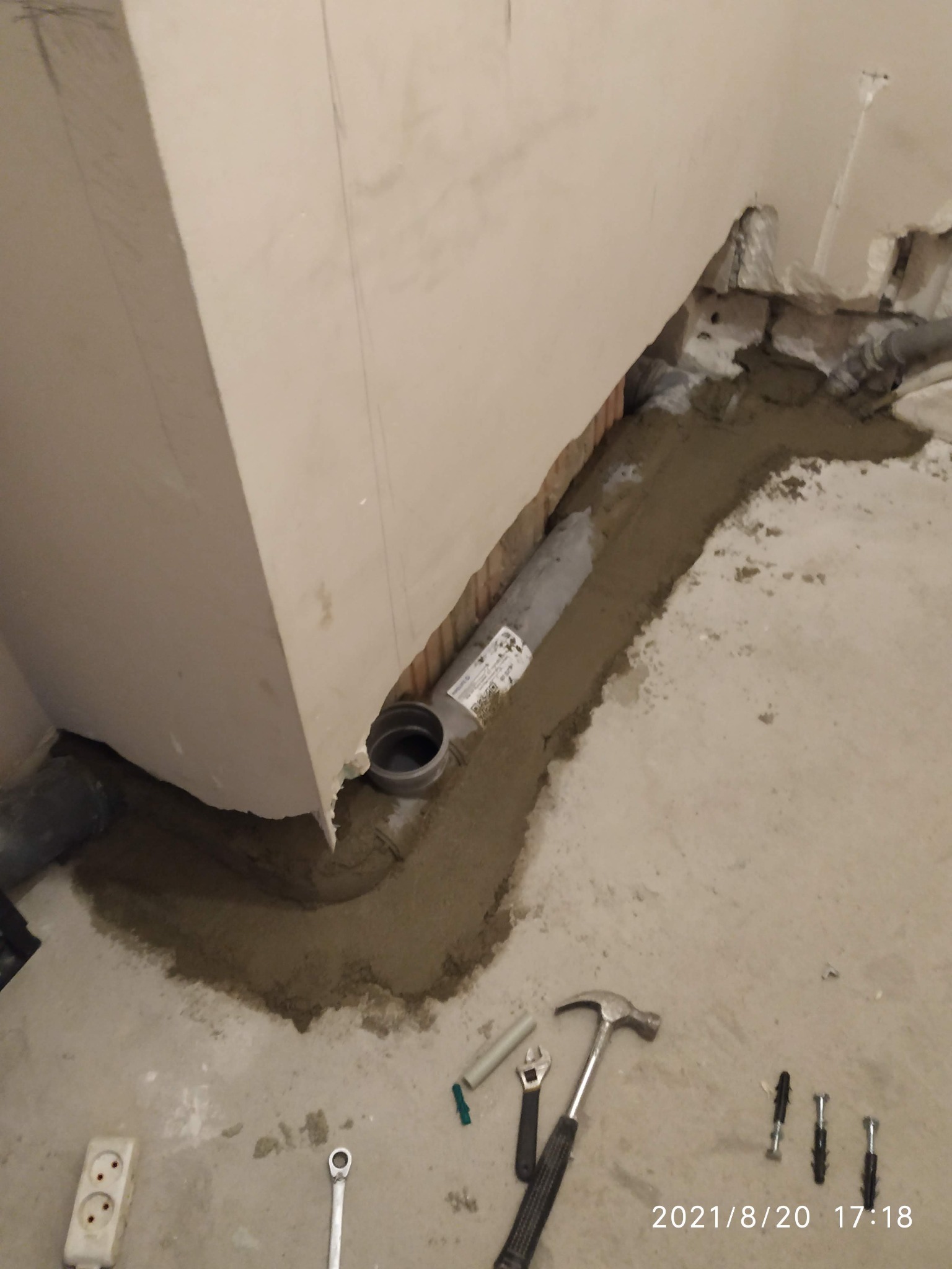 Szara rura kanalizacyjna w wyżłobieniu w posadzce, zalana świeżym cementem, widoczne narzędzia: młotek, klucz nastawny, klucz oczkowy, kołki rozporowe, gniazdko elektryczne w tle ściana w trakcie...