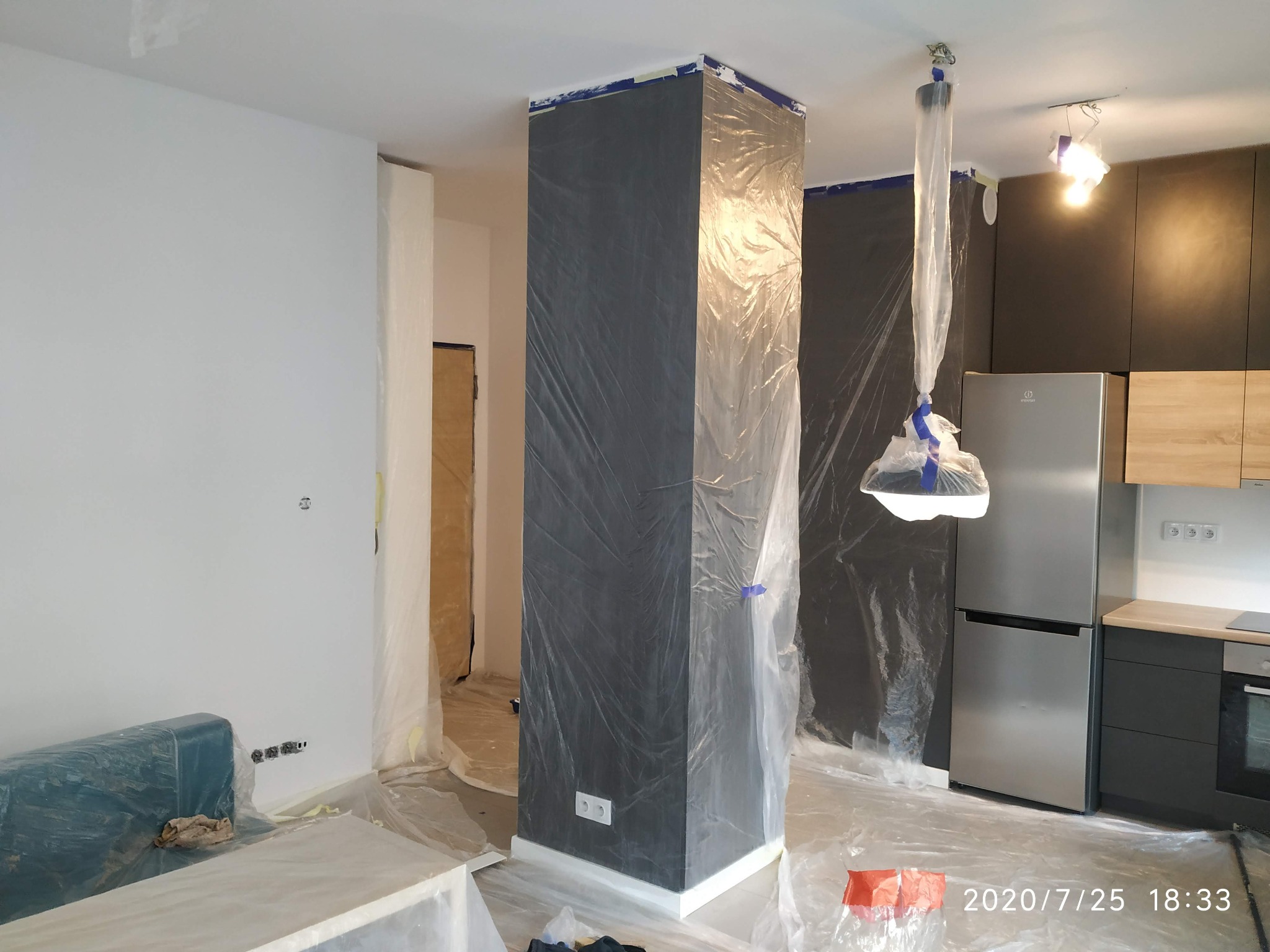 Pokój w trakcie zabezpieczania folią malarską przed malowaniem ścian i kolumny, widoczna kuchnia z zabudową i sprzętem AGD również osłonięta folią, gniazdka elektryczne zabezpieczone taśmą.