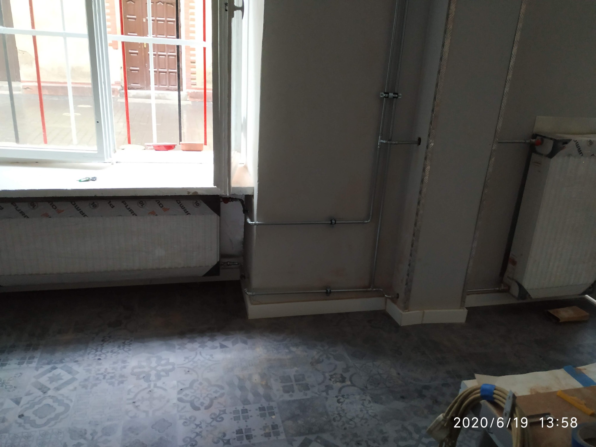 Nowe, wzorzyste płytki podłogowe w szarościach, zainstalowane rury grzewcze przy ścianie oraz ocieplony parapet okienny w trakcie prac wykończeniowych.