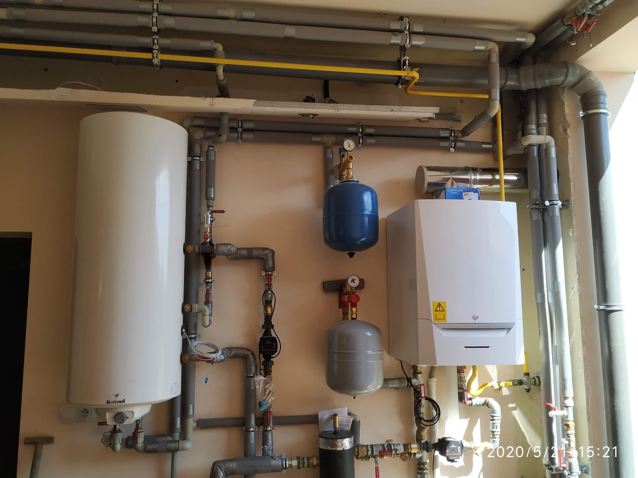 Kaskada instalacji centralnego ogrzewania z kotłem gazowym De Dietrich, zasobnikiem Galmet, pompami obiegowymi i zbiornikami wyrównawczymi, widoczne rury instalacyjne z zaworami i manometrami.