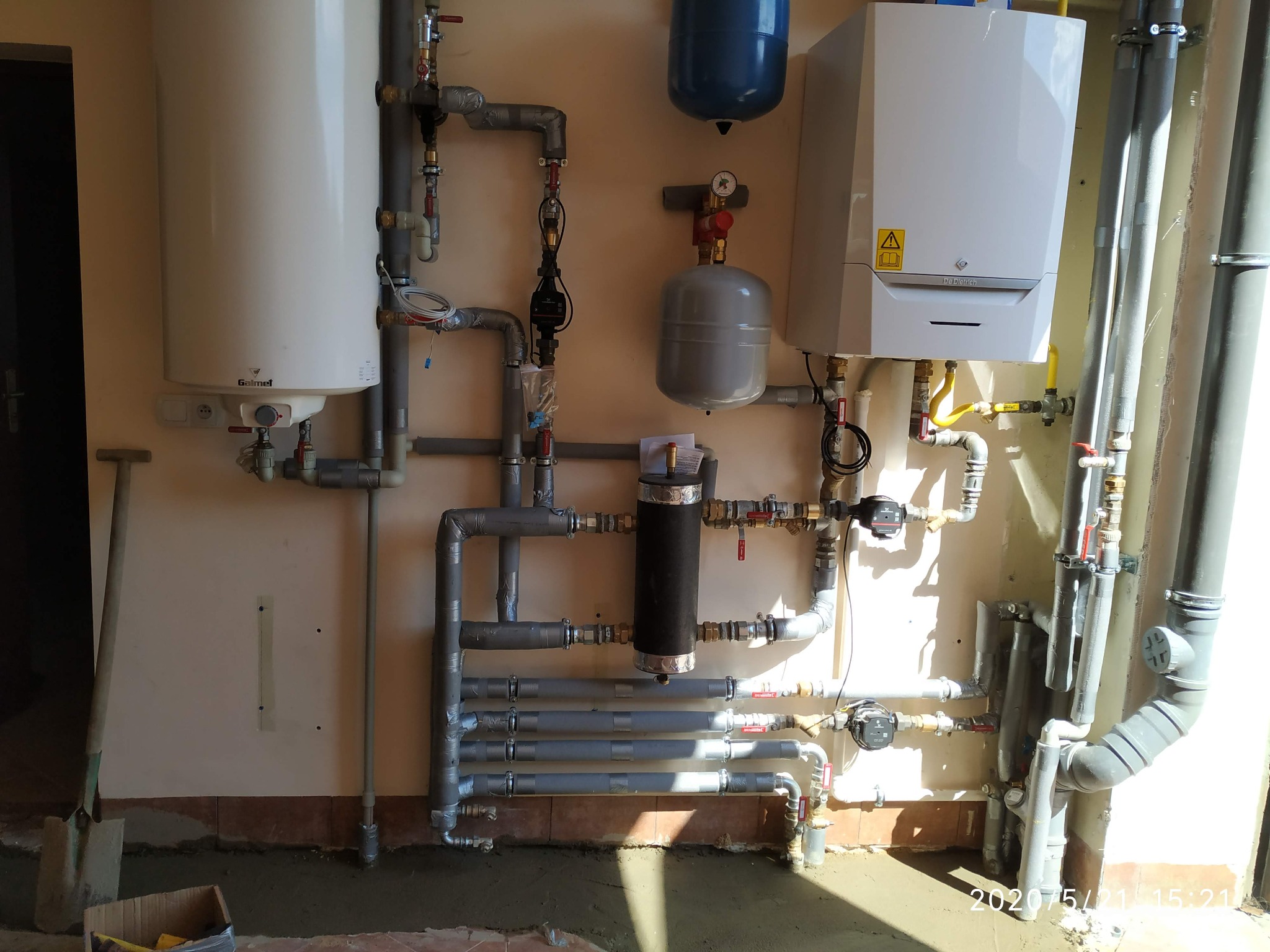 Kompleksowa instalacja grzewcza z kotłem De Dietrich, zasobnikiem Galmet i licznymi rurami, widoczny etap montażu z resztkami zaprawy na podłodze i łopatą w tle.