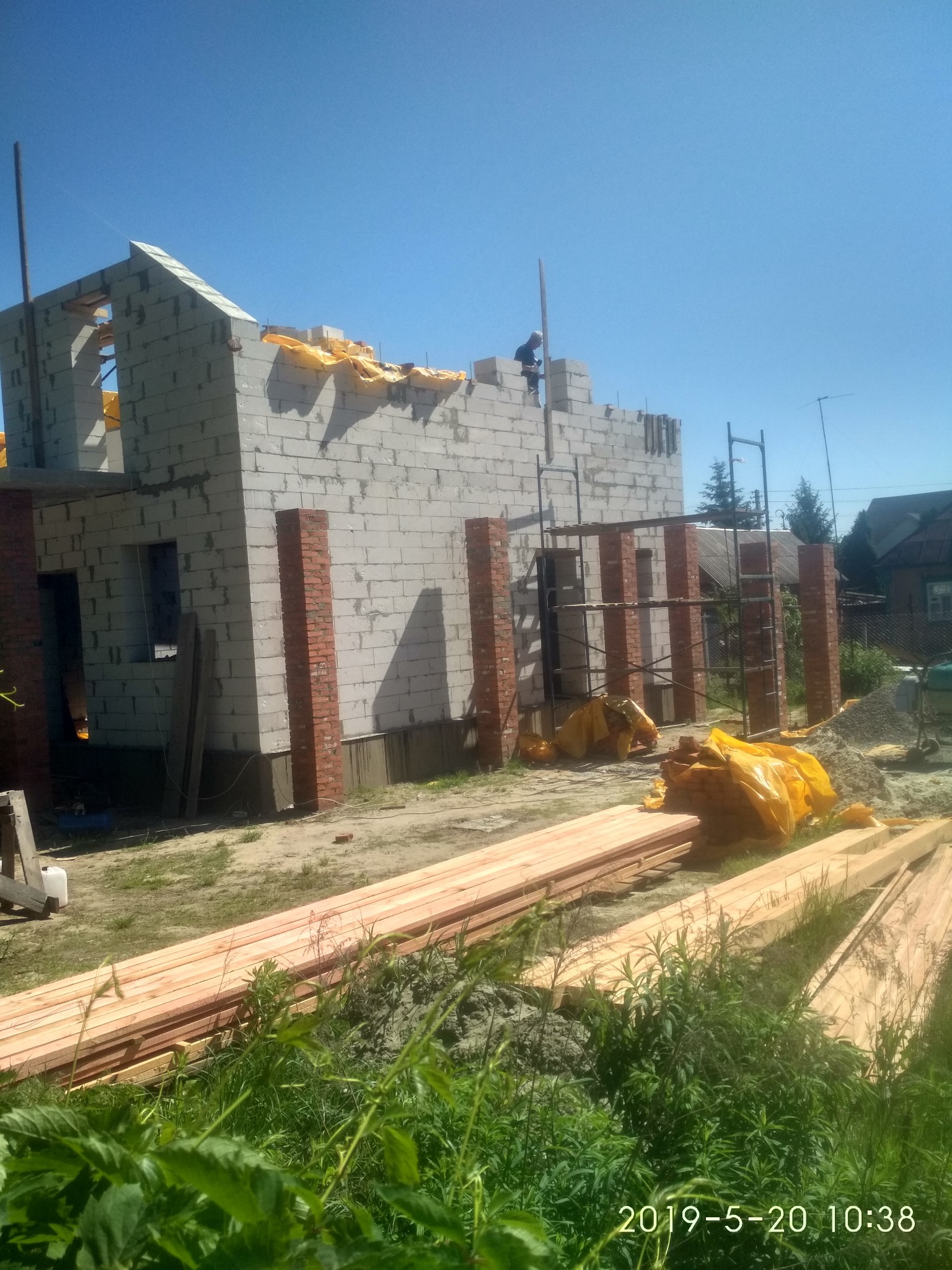 Budowa domu w Łodzi: surowy stan z białych bloczków, częściowo obłożony czerwoną cegłą, drewniane stemple, widoczne rusztowania i składowane deski na tle błękitnego nieba.