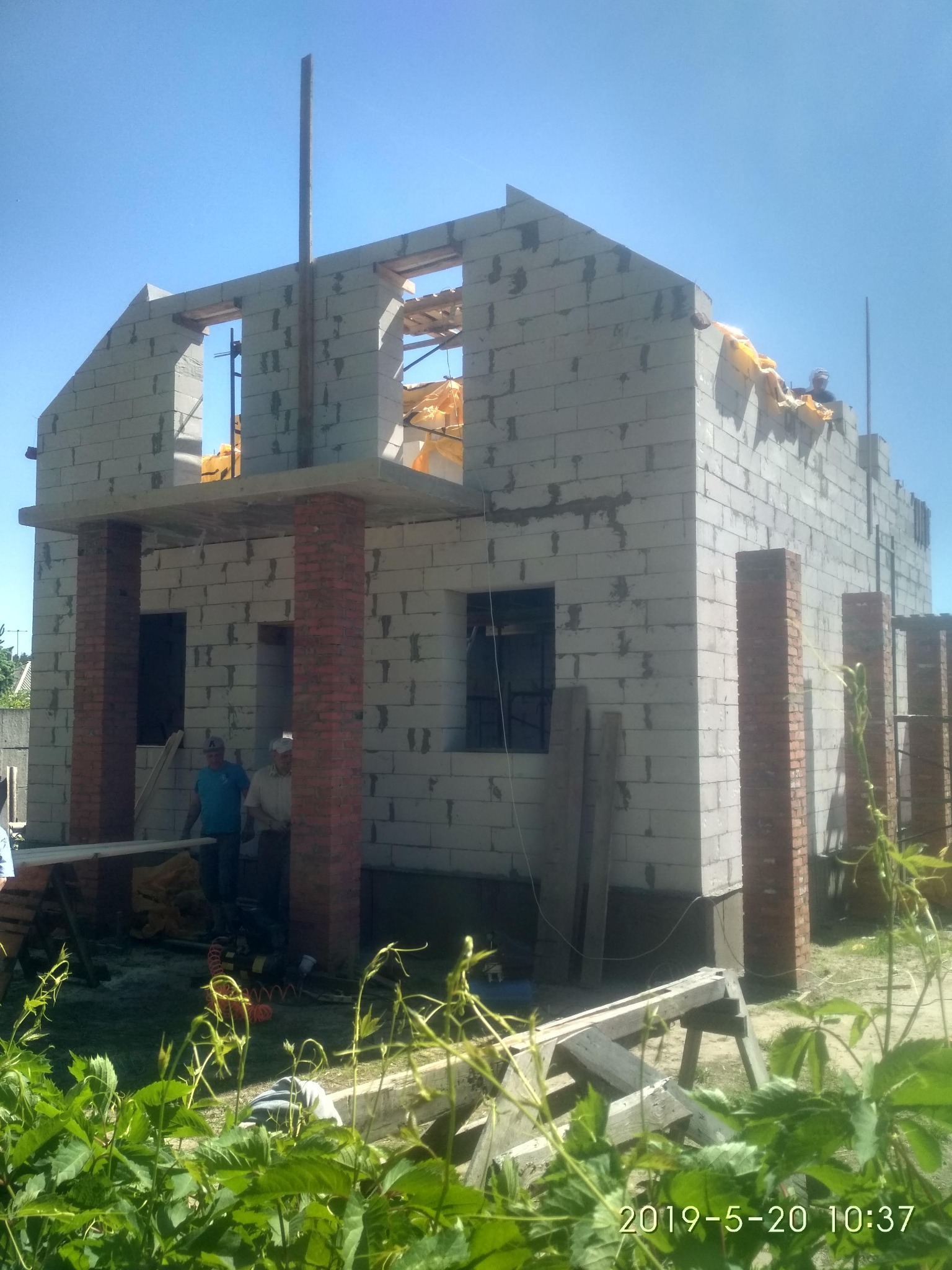 Surowy stan budynku w trakcie wznoszenia, ściany z betonu komórkowego i ceglane filary, widoczni dwaj pracownicy oraz narzędzia budowlane na tle zieleni.