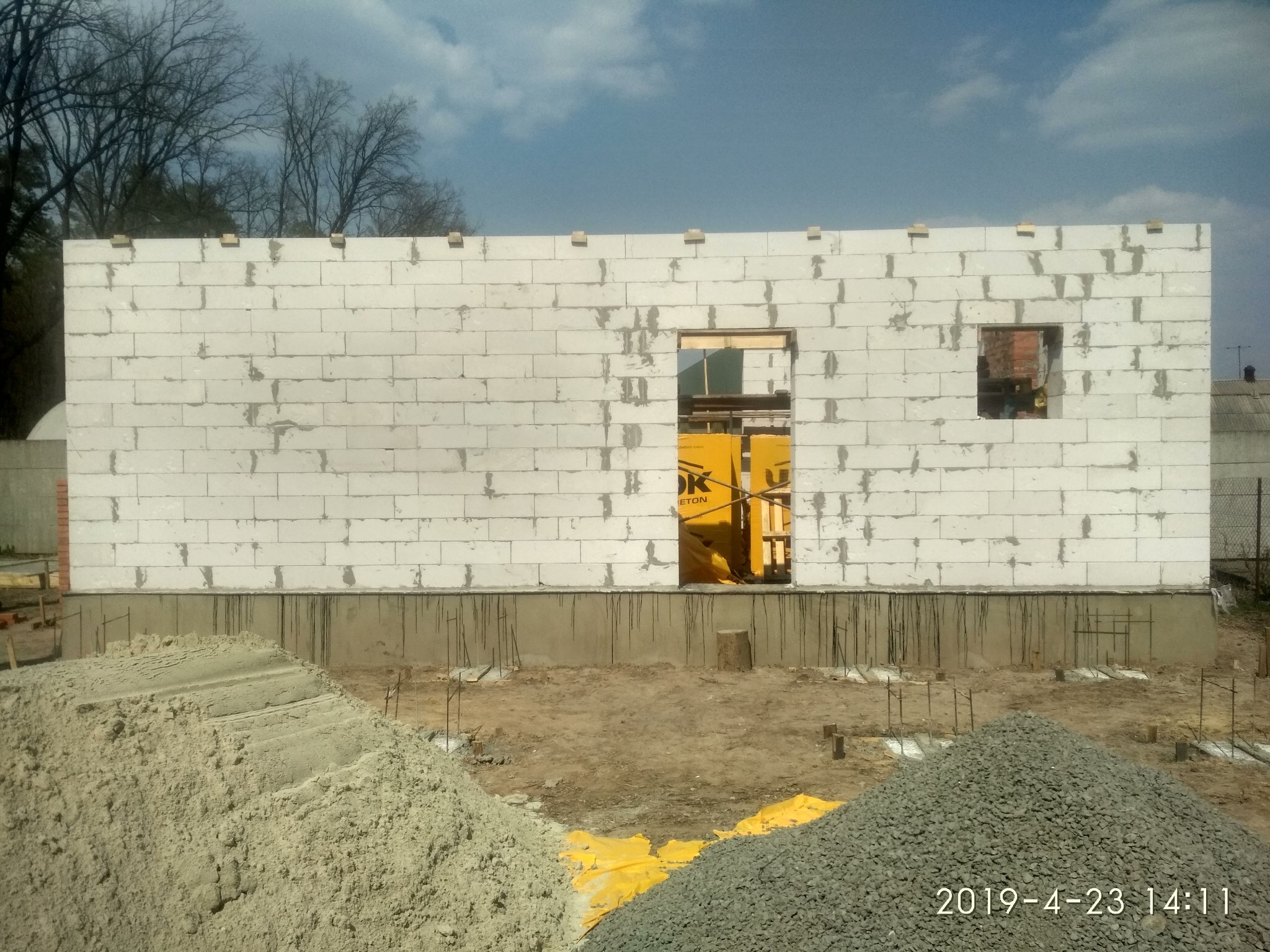 Wznoszenie ściany z białych bloczków betonowych na fundamencie, widoczne otwory okienne i drzwiowe zabezpieczone folią, na pierwszym planie hałdy piasku i żwiru.