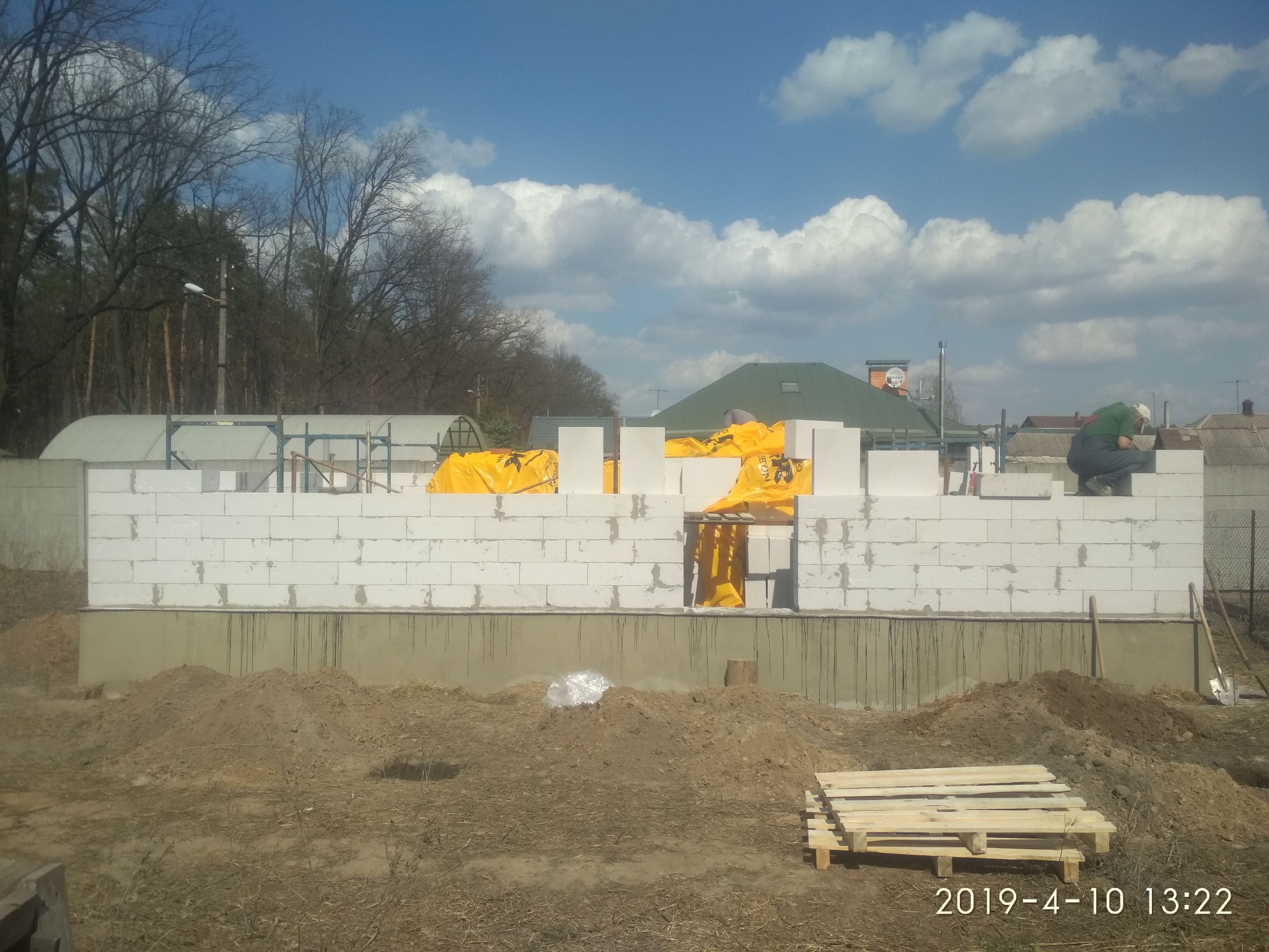 Wznoszenie muru z białych bloczków, częściowo zabezpieczonych żółtą folią, na fundamencie; w tle widoczny pracownik w zielonej koszuli, drzewa i zabudowania mieszkalne.