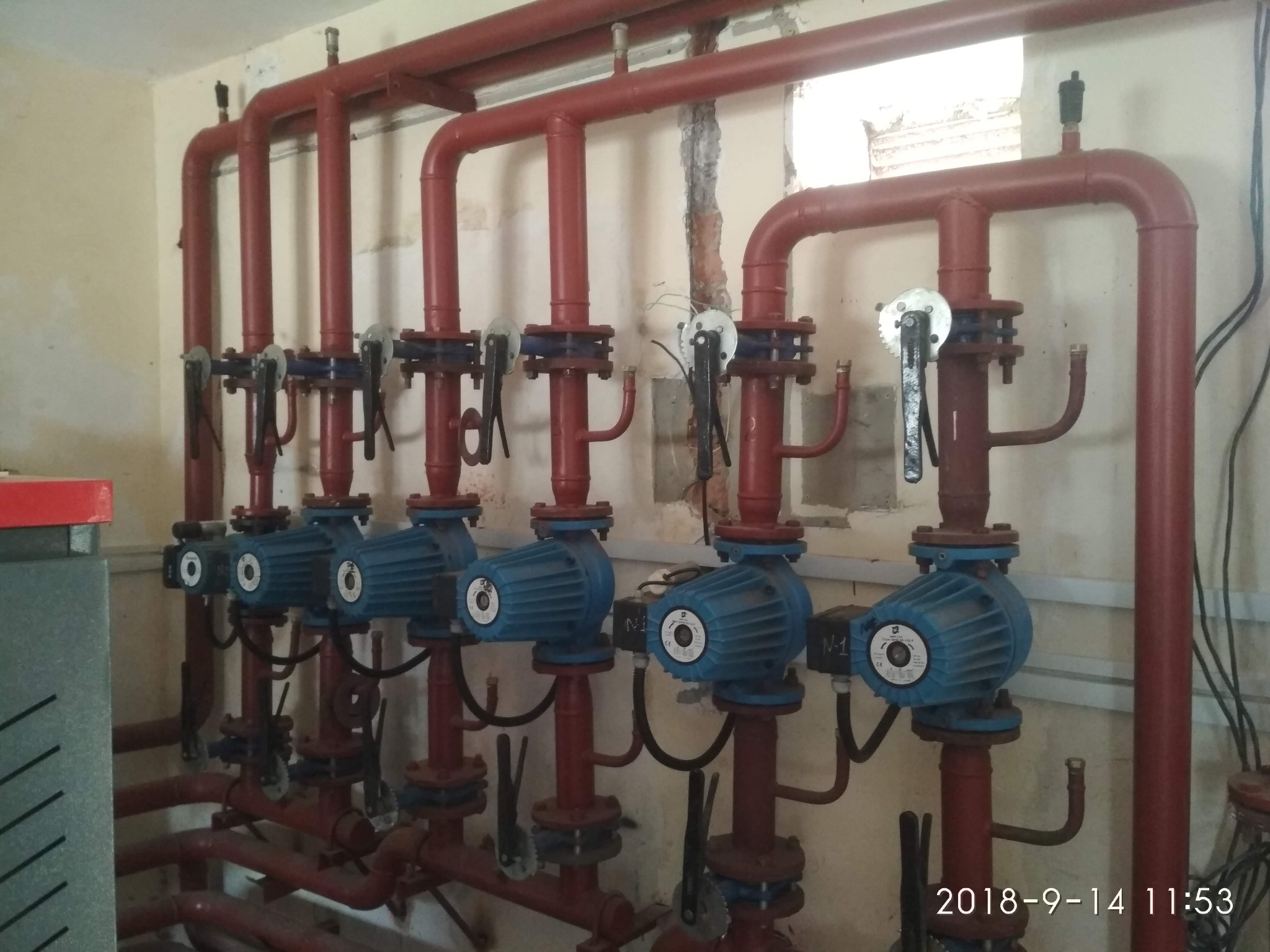 Szereg sześciu niebieskich pomp obiegowych zamontowanych na czerwonych rurach w kotłowni, widoczne zawory i instalacja hydrauliczna, ściany w tle z widocznymi śladami napraw.