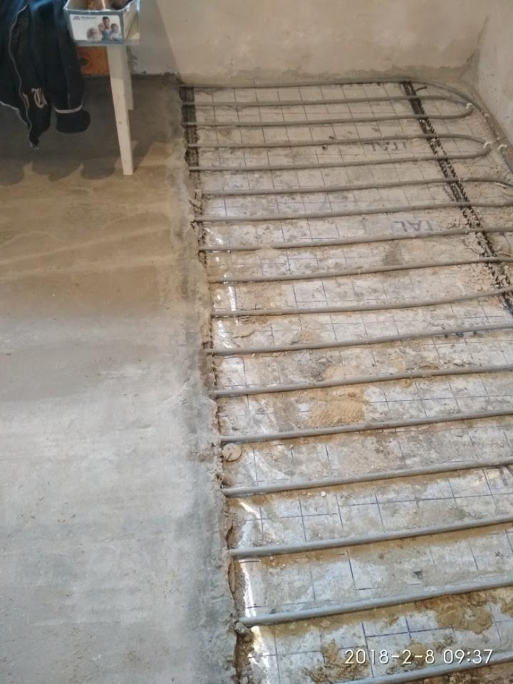 Montaż rur ogrzewania podłogowego na siatce w surowym pomieszczeniu, widoczne niedoskonałości podłoża i data na zdjęciu.