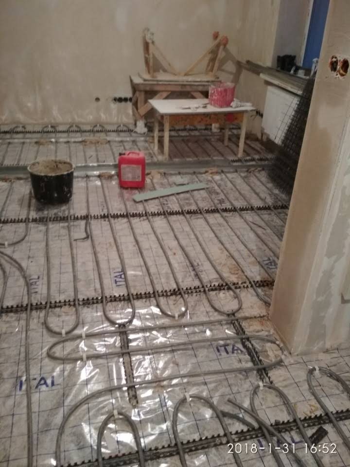 Montaż ogrzewania podłogowego: rury grzewcze rozłożone na folii z nadrukiem 'ITAL' na podłożu przed wylaniem wylewki, widoczny pojemnik z zaprawą i czerwony kanister w tle, surowe ściany.