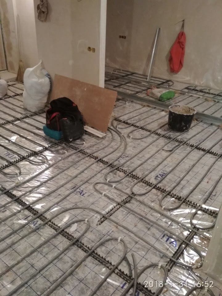 Montaż ogrzewania podłogowego: rury grzewcze ułożone na folii z nadrukiem, widoczne torby budowlane, wiadro i narzędzia w surowym wnętrzu.