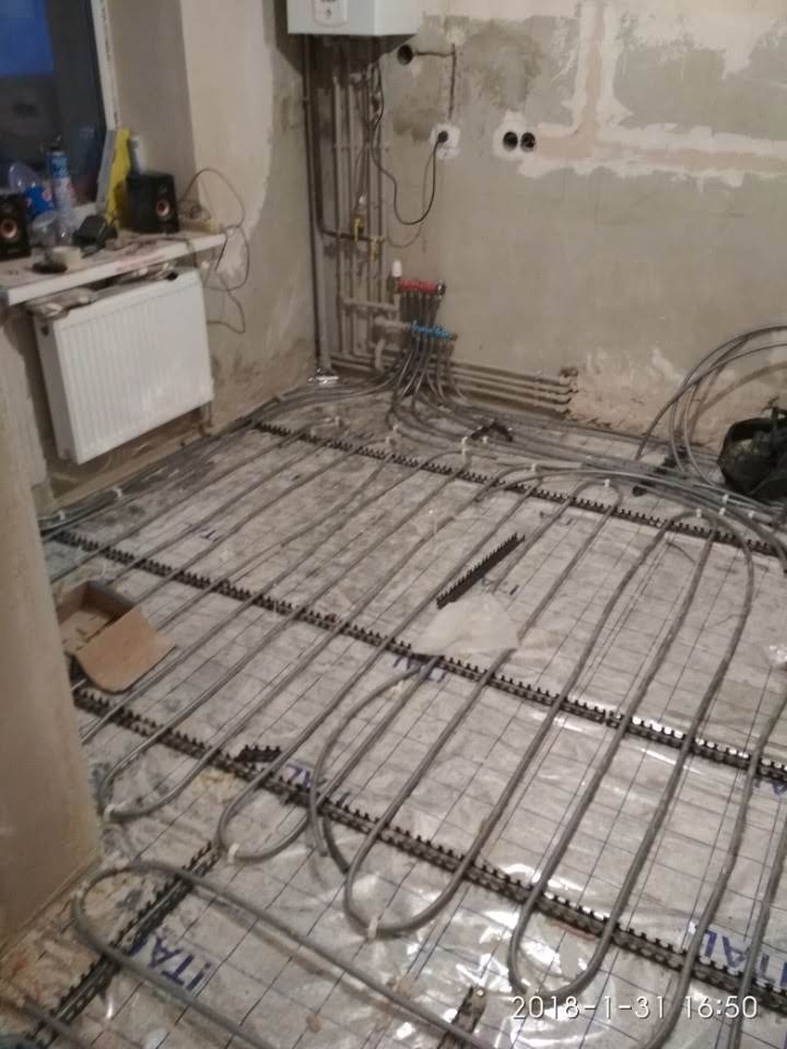Montaż ogrzewania podłogowego: rury grzewcze ułożone na siatce, widoczne elementy instalacji hydraulicznej przy ścianie, surowy stan wykończenia wnętrza.