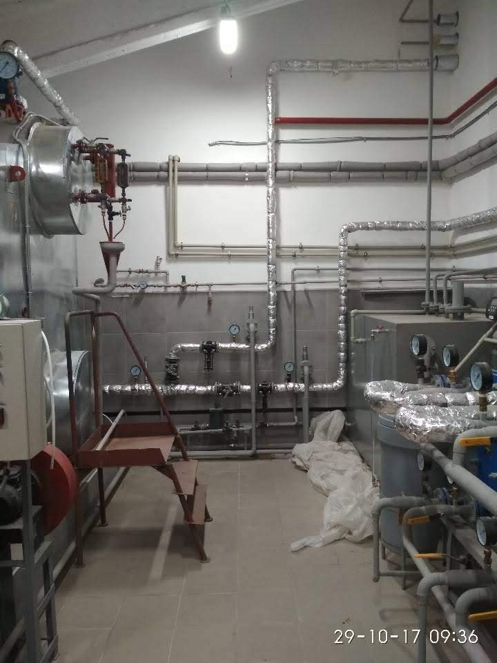 Kotłownia z licznymi rurami, zaworami i manometrami, częściowo zaizolowanymi folią aluminiową, widoczne schodki do obsługi urządzeń, podłoga wyłożona płytkami ceramicznymi.