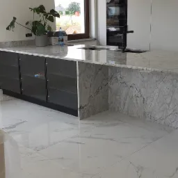 Nowoczesna kuchnia z marmurową wyspą, czarne szafki z przeszklonymi frontami, białe płytki podłogowe z szarym wzorem, okno z brązową ramą i rośliny doniczkowe na blacie.