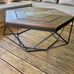 Agnieszka Bielicka Agni Metal - Stylowy stolik kawowy z drewnianym blatem o heksagonalnym kształcie i metalową, geometryczną podstawą. Ustawiony na drewnianym tarasie, w tle beżowa sofa z poduszkami.