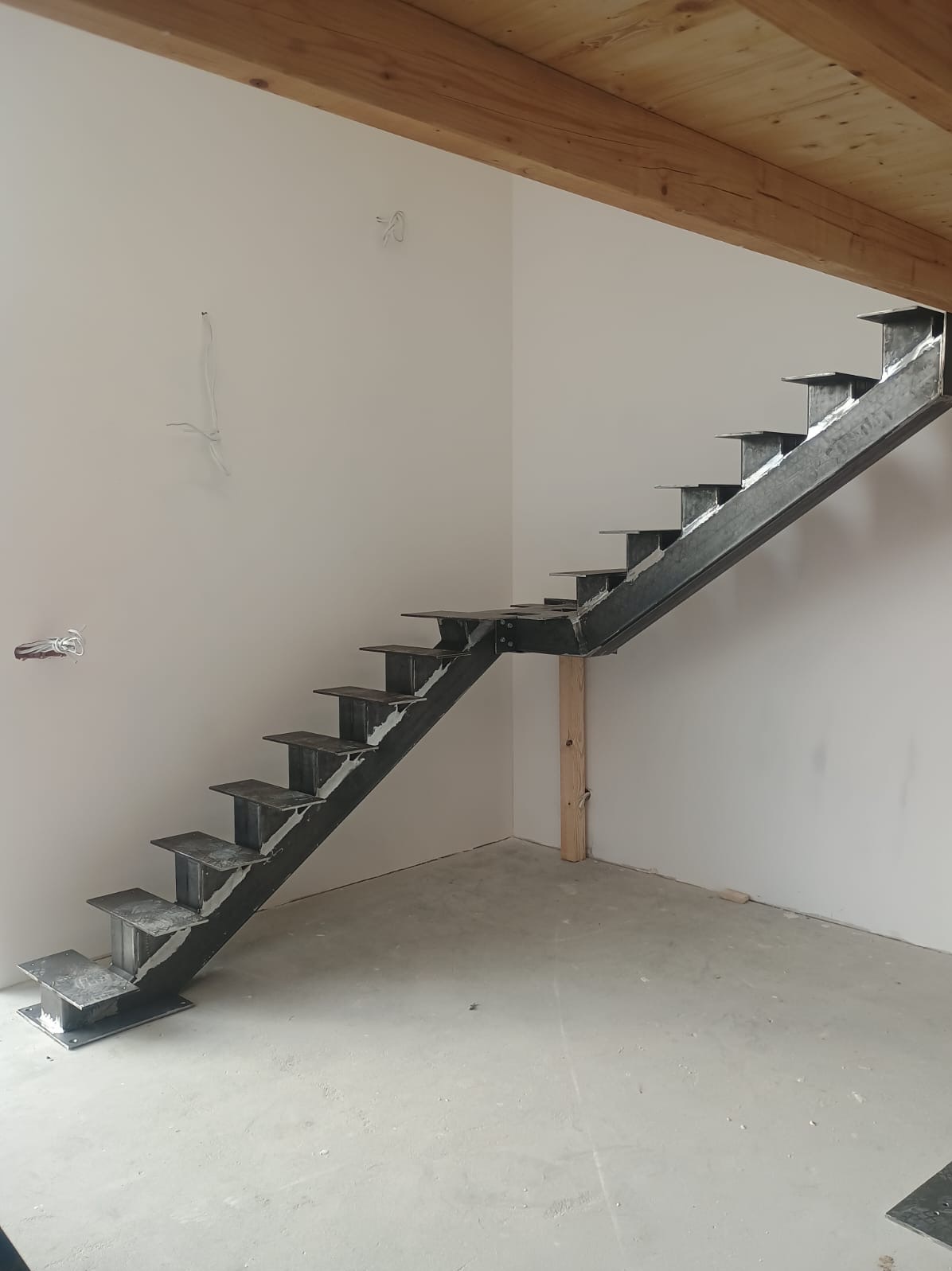 Surowe, spawane schody metalowe w jasnym wnętrzu, wsparte na drewnianym słupku. Widoczne ślady spawania i niedokończone wykończenie. Minimalistyczny design, surowy stan.