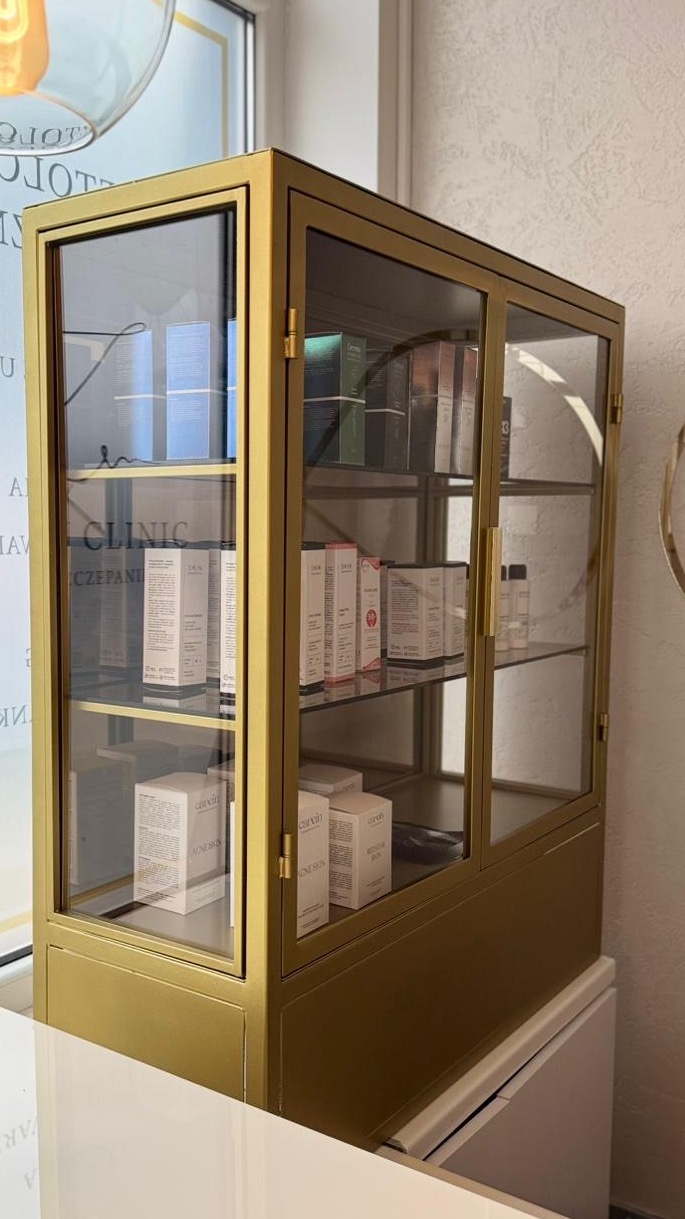 Złota, przeszklona witryna z kosmetykami w salonie. Widoczne półki z produktami do pielęgnacji skóry. Minimalistyczny design, jasne wnętrze, elegancki wygląd.