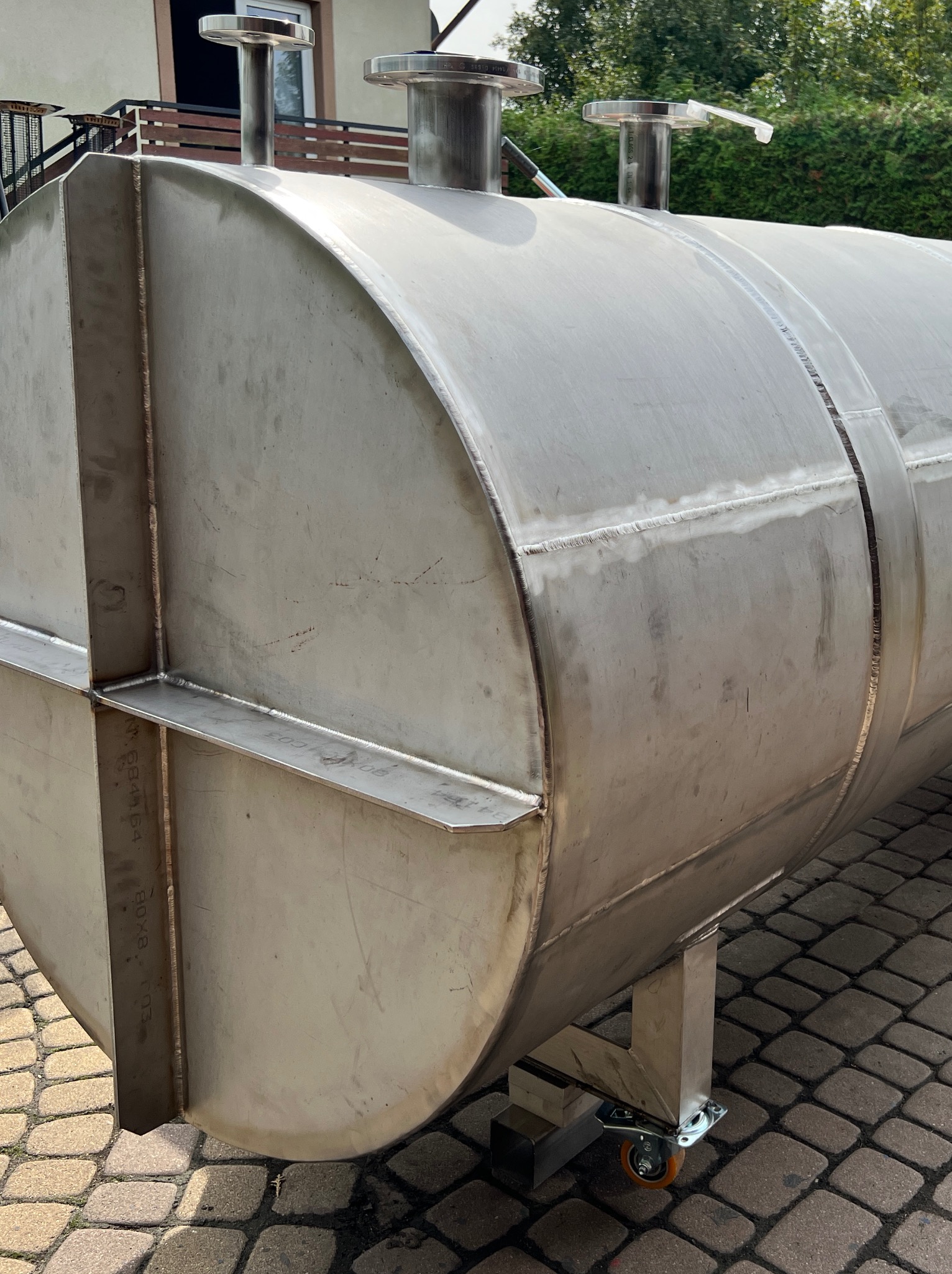 Zbliżenie na spawany, cylindryczny zbiornik ze stali nierdzewnej na kółkach, z widocznymi spawami i elementami mocującymi. Tło stanowi kostka brukowa i zieleń.