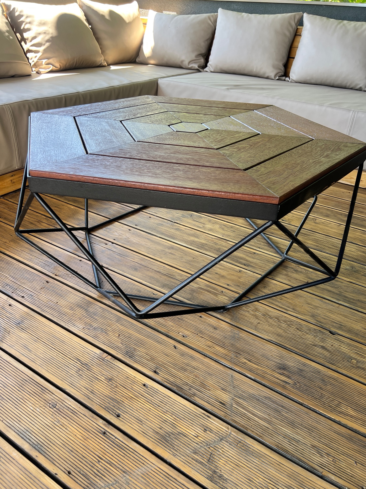 Stylowy stolik kawowy z drewnianym blatem o heksagonalnym kształcie i metalową, geometryczną podstawą. Ustawiony na drewnianym tarasie, w tle beżowa sofa z poduszkami.