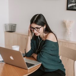 DirectAcademy Aleksandra Siedlecka - Młoda kobieta w okularach i zielonym swetrze, z białymi słuchawkami, uśmiecha się, patrząc na srebrnego laptopa na drewnianym stole, prawdopodobnie podczas lekcji online. W tle jasna ściana...
