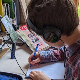 DirectAcademy Aleksandra Siedlecka - Uczeń w słuchawkach Sony, piszący w zeszycie w kratkę, z otwartym podręcznikiem z sekcjami 'Speaking' i 'Reading' oraz tabletem na biurku, w domowym zaciszu.