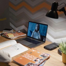 DirectAcademy Aleksandra Siedlecka - Mały chłopiec uczy się zdalnie, oglądając kobietę na ekranie laptopa. Na biurku otwarte książki, w tym 'ENTERPRISE 2', i lampka. Ściana z wzorem chevron.