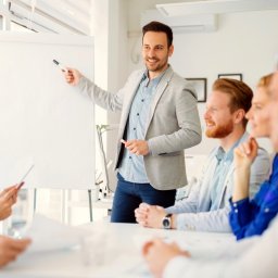 DirectAcademy Aleksandra Siedlecka - Uśmiechnięty prezenter w szarej marynarce wskazuje czarnym markerem na pustą białą tablicę, prowadząc spotkanie dla grupy słuchaczy siedzących przy stole w jasnym biurze.