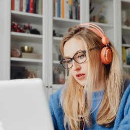 DirectAcademy Aleksandra Siedlecka - Blondynka w okularach i pomarańczowych słuchawkach na uszach, skupiona na ekranie srebrnego laptopa, z białym regałem pełnym kolorowych książek w tle.