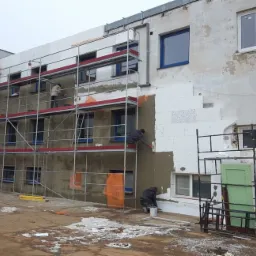 Ocieplanie elewacji budynku z wykorzystaniem rusztowania, widoczne fragmenty starej elewacji i nowe okna z niebieską ramą.