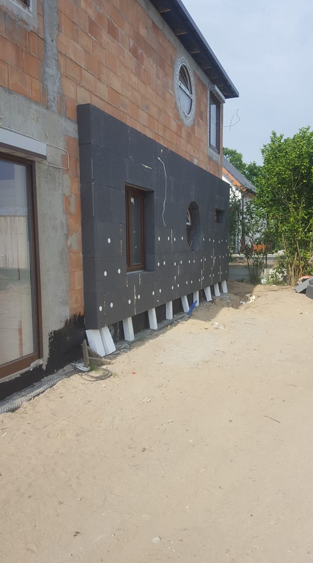 Częściowo ocieplona ściana budynku z cegły z okrągłym oknem, widoczne białe kołki mocujące i czarna powłoka hydroizolacyjna u dołu, przygotowana do dalszych prac.