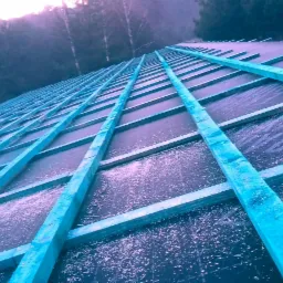 Konstrukcja dachu z impregnowanych na niebiesko łat, pokryta czarną membraną dachową z widocznym nadrukiem, na tle zalesionego terenu w pochmurny dzień.