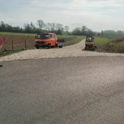 Nowa asfaltowa droga z pomarańczową ciężarówką i koparką w tle oraz pracownikiem w kamizelce odblaskowej z boku. Widoczny znak drogowy.