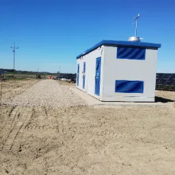 Szara, modułowa stacja transformatorowa z niebieskimi akcentami na terenie budowy farmy fotowoltaicznej. Utwardzony teren wokół budynku.