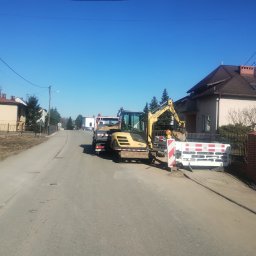 Bamber FV Kamil Kaczmarski - Żółta koparka Yanmar SV26 na ulicy, obok bariera ochronna. W tle domy i błękitne niebo. Prace ziemne w toku, widoczny wykop.