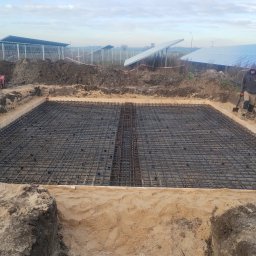 Bamber FV Kamil Kaczmarski - Zbrojenie fundamentu pod panele słoneczne, widoczne szalunki i pracownicy na budowie. W tle panele słoneczne i generator prądu.