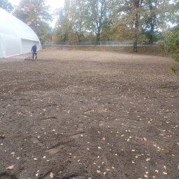 Bamber FV Kamil Kaczmarski - Pracownik wyrównuje teren walcem ogrodowym na działce z brązową ziemią i opadłymi liśćmi, w tle biały namiot i drzewa.