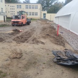 Bamber FV Kamil Kaczmarski - Pomarańczowa ciężarówka z koparką na placu budowy, widoczne ślady opon na ziemi, budynki w tle i fragment hali namiotowej. Prace ziemne w toku.