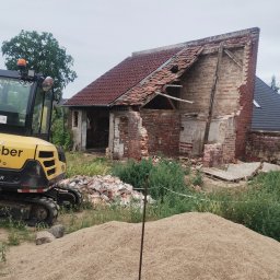 Bamber FV Kamil Kaczmarski - Częściowo rozebrany budynek z czerwonej cegły, gruz i żółta koparka w trawie. Widoczne uszkodzenia dachu i ścian. Etap rozbiórki z bliska.