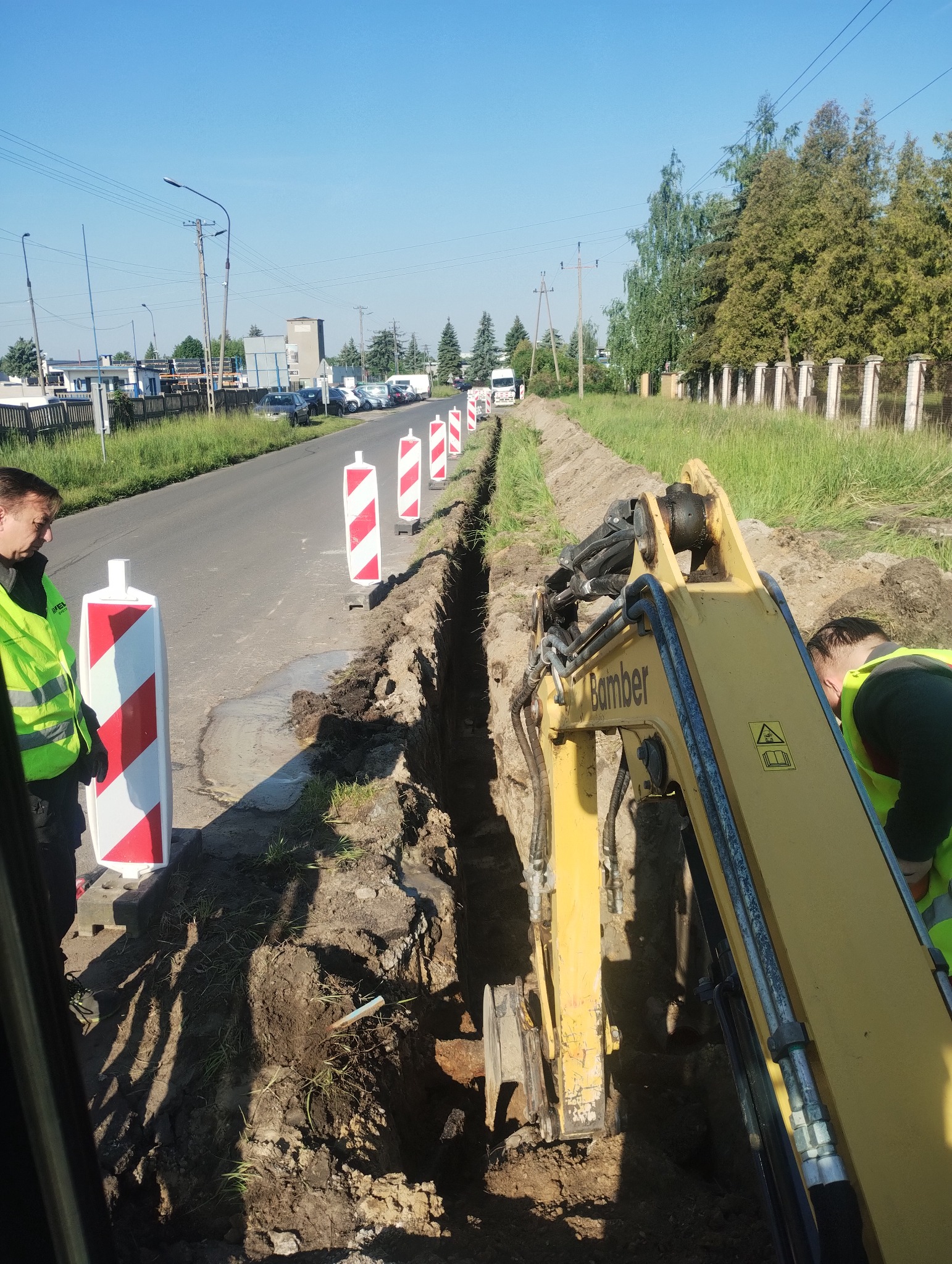 Wykop liniowy pod instalacje, zabezpieczony słupkami ostrzegawczymi. Żółta koparka BamBer w trakcie pracy, dwóch pracowników w odblaskowych kamizelkach nadzoruje wykop.