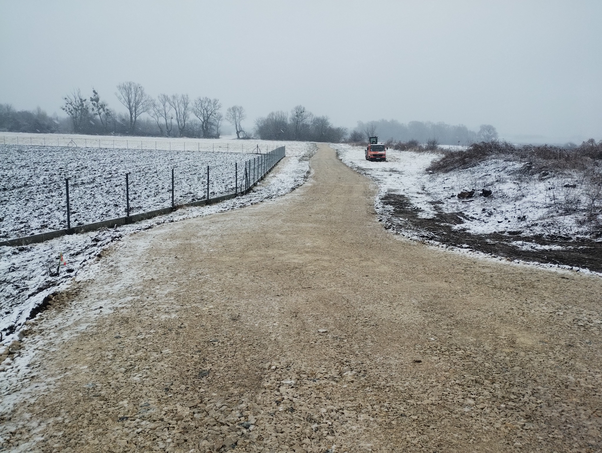 Utwardzona, szutrowa droga w zimowej scenerii, z częściowo ośnieżonym polem po lewej i czerwonym pojazdem w oddali. Metalowe ogrodzenie wzdłuż drogi.