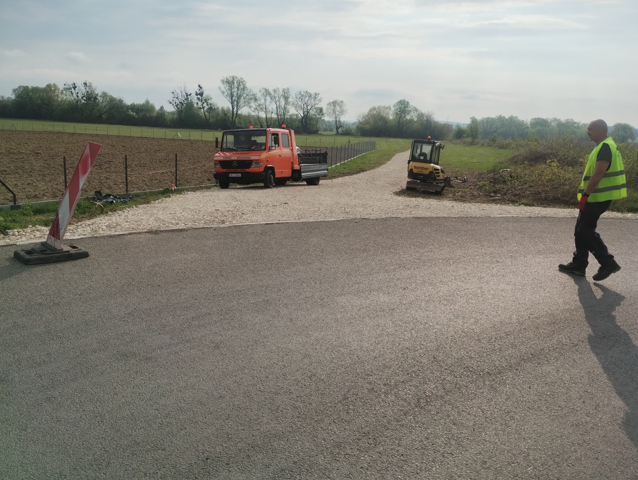 Nowa asfaltowa droga z pomarańczową ciężarówką i koparką w tle oraz pracownikiem w kamizelce odblaskowej z boku. Widoczny znak drogowy.