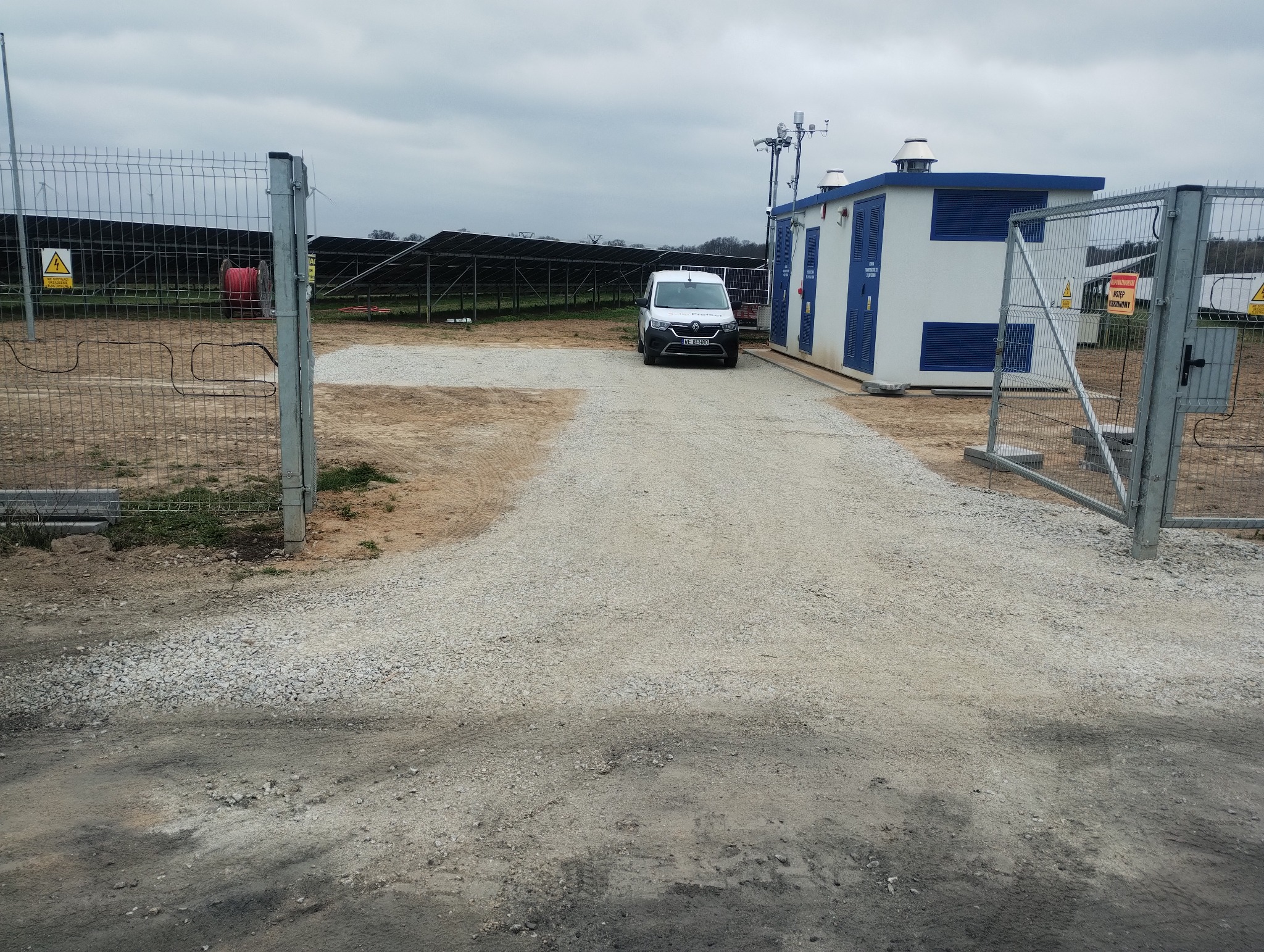 Utwardzona żwirowa droga w kierunku budynku stacji transformatorowej i paneli słonecznych. Otwarta brama wjazdowa z siatki, biały samochód dostawczy w tle.