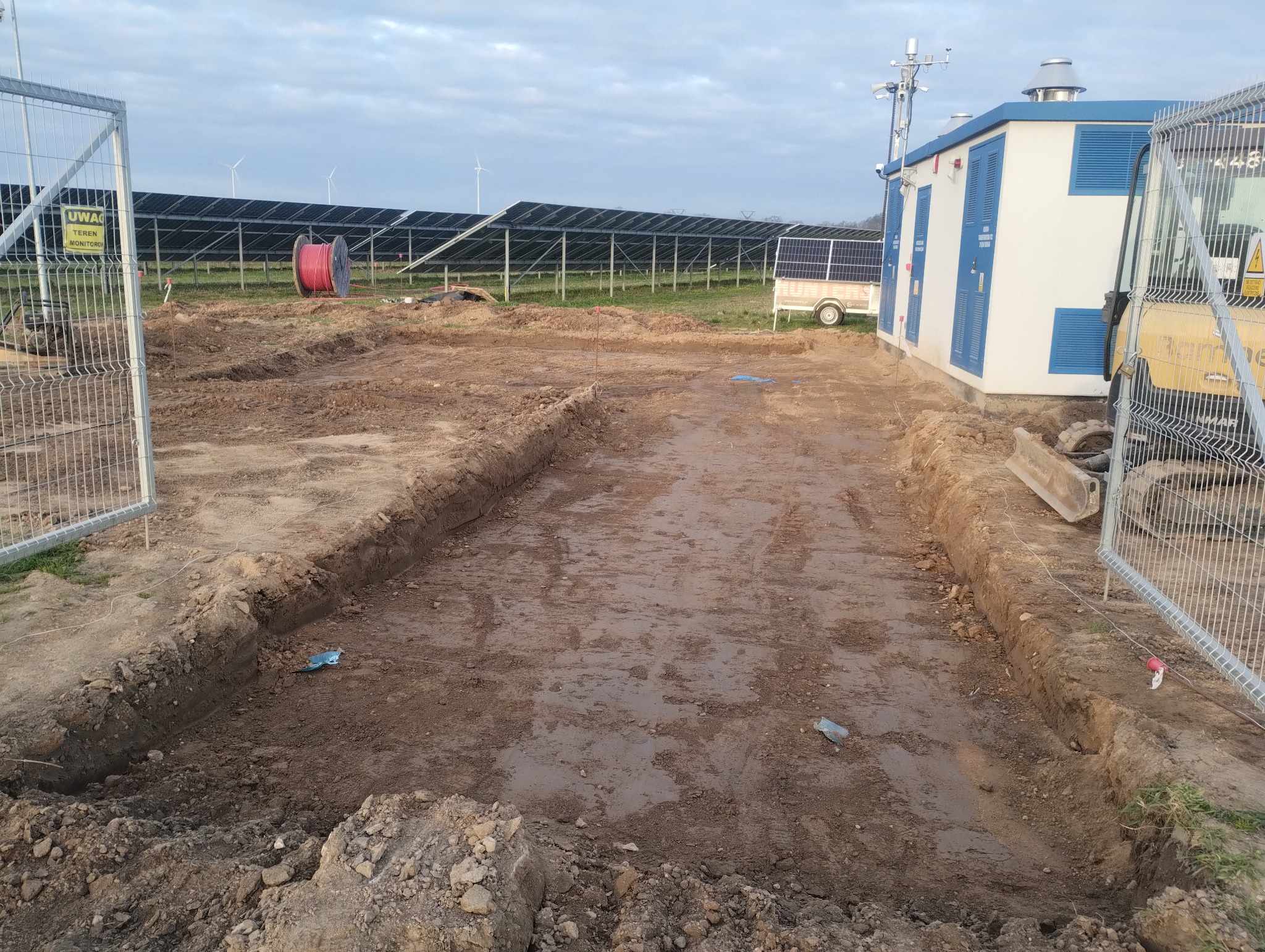 Wykop liniowy w ziemi, prawdopodobnie pod instalacje, z widocznymi panelami słonecznymi w tle. Na obrzeżach wykopu widoczny sprzęt budowlany i ogrodzenie.