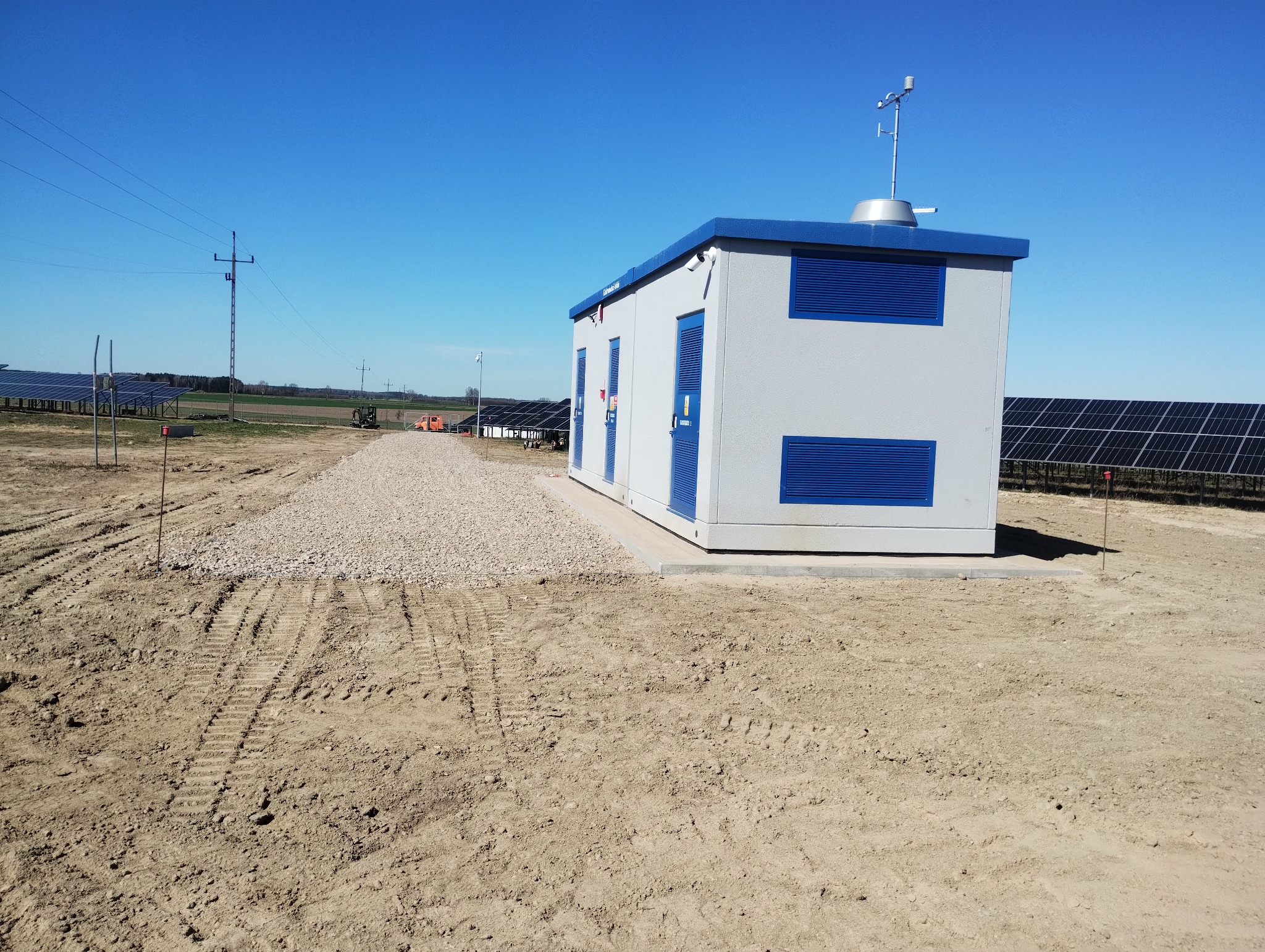 Szara, modułowa stacja transformatorowa z niebieskimi akcentami na terenie budowy farmy fotowoltaicznej. Utwardzony teren wokół budynku.