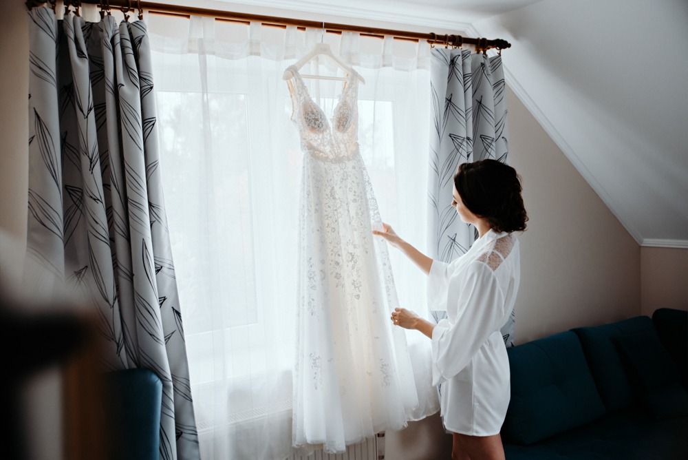 Panna młoda w białym szlafroku ogląda wiszącą suknię ślubną z koronkowymi detalami na tle okna z wzorzystymi zasłonami.