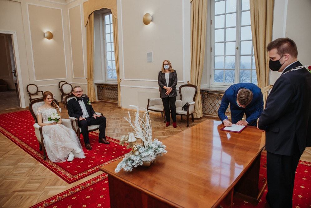 Para młoda siedzi podczas ceremonii ślubnej, urzędnik w masce nadzoruje składanie podpisu przez świadka na drewnianym stole z dekoracją.
